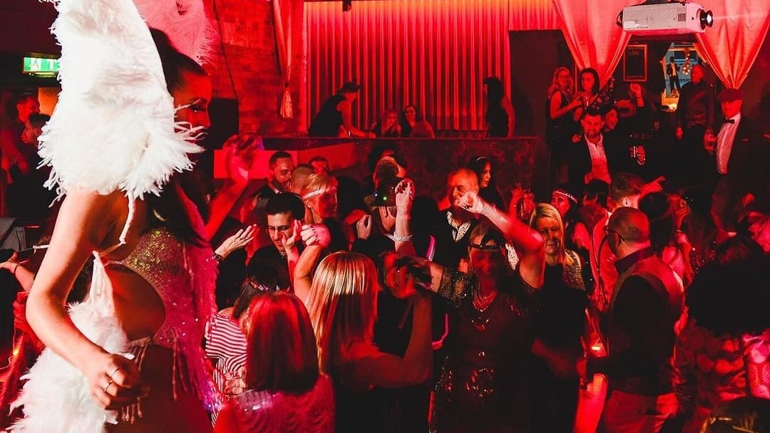A lively nightclub scene at Canvas Bournemouth with a vibrant crowd dancing under red lighting, featuring a performer in an elaborate feather costume interacting with guests.